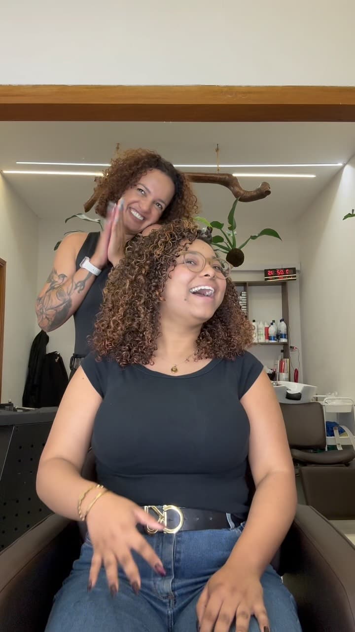 Cabeleireira finalizando os cachos de uma cliente que ri na cadeira