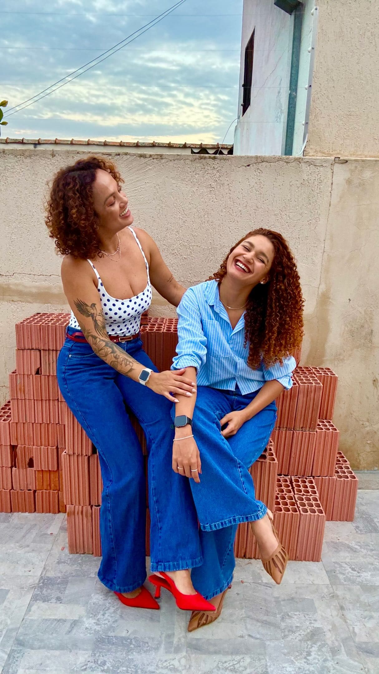 As duas profissionais do salão, de cachos ruivos, rindo juntas sentadas sobre tijolos
