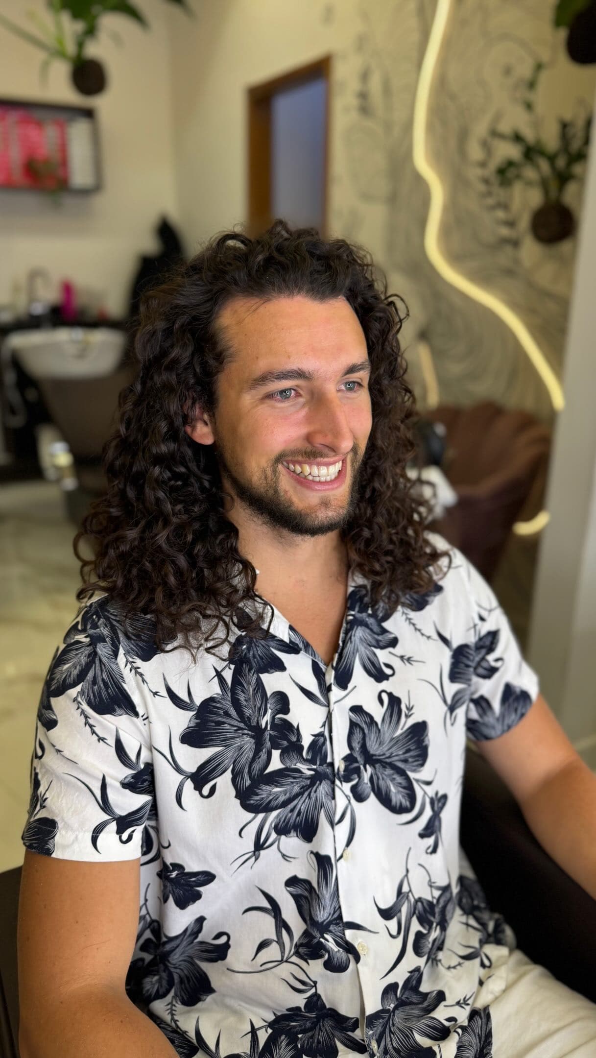 Rapaz de cachos longos sorrindo na cadeira, em frente ao mural do salão